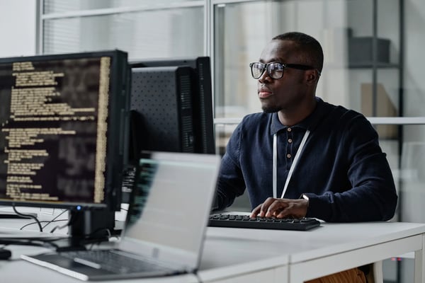 homem sentado em frente ao computador. Atrás de sua tela, outro monitor aparece mostrando a tela para a câmera.