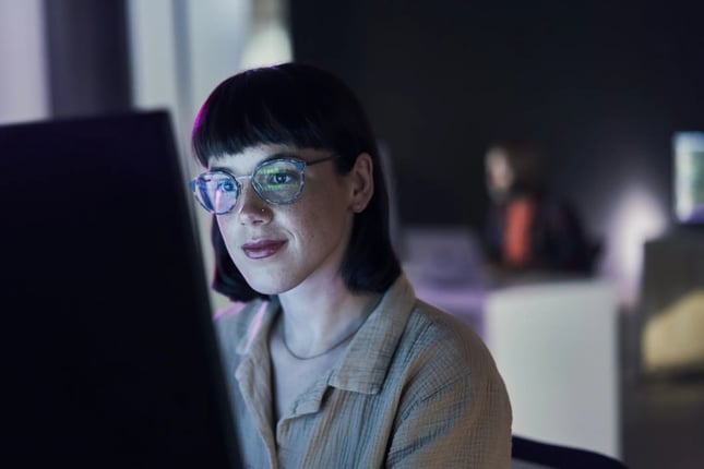 Uma mulher de frente para a tela de um computador. Em seu óculos de grau, reflete-se um código-fonte, sugerindo que ela atua no setor de tecnologia. 