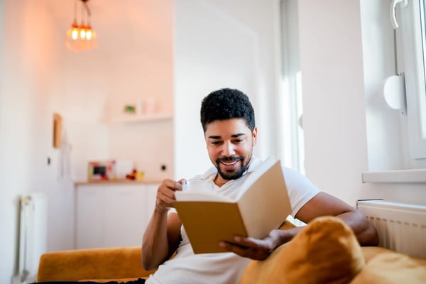 Um homem jovem e negro está sentado no sofá, sorrindo, enquanto lê um livro. O tema do artigo é livros sobre liderança.