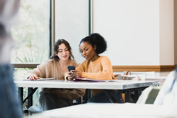 A imagem traz duas adolescentes multiétnicas que olham para o telefone inteligente enquanto palestras de professor irreconhecíveis. O tema do artigo é SXSW 2024.
