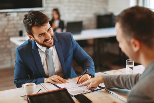 A imagem apresenta dois executivos jovens e brancos que estão em um escritório vestindo roupas formais e apertando ambas as mãos. O tema do artigo é captação de recursos para startups.