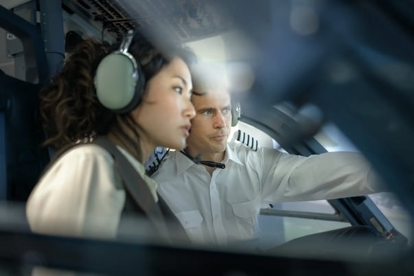 Mulher e homem em uma cabine de avião. Os dois vestem roupas características da aviação e fones de ouvido abafadores.