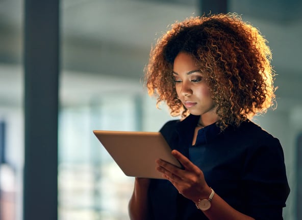 a imagem mostra uma mulher em pé, segurando um tablet com as duas mãos. Ela está em um escritório, com a tela de frente para seu rosto.
