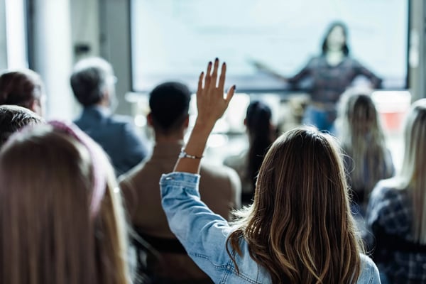 A imagem apresenta uma mulher de costas em uma multidão que levanta uma das mãos para fazer uma pergunta. O tema do artigo é curva de inovação.