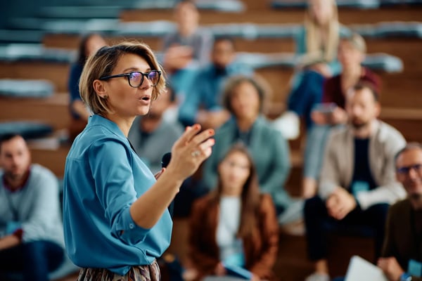 A imagem apresenta uma palestrante mulher fazendo uma apresentação durante seminário de negócios no centro de convenções. O tema do artigo é empreendedorismo corporativo.
