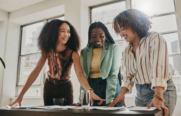 Um grupo de três mulheres jovens, sorridentes e negras, estão em frente a uma mesa de reuniões, como a trabalhar em um projeto. Elas vestem roupas casuais e o clima é de descontração. O tema do artigo é sobre metodologias de gestão de metas e como usar nas organizações.
