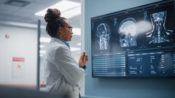 A imagem apresenta uma médica em frente a um painél digital, avaliando gráficos e imagens digitais de um paciente. A mulher é jovem e parda, e usa jaleco branco. O tema do artigo é inteligência artificial na saúde.