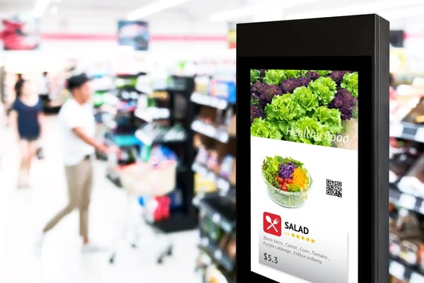 A imagem apresenta um totem digital, em cuja tela aparece uma propaganda em inglês vendendo uma salada, como se fosse uma oferta do dia do supermercado. Ao fundo, pessoas estão num supermercado, fazendo compras. O tema do artigo é inteligência artificial no varejo.