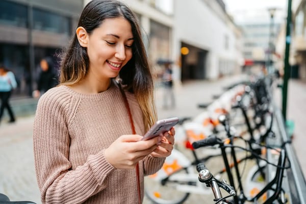 Na imagem, uma mulher jovem e branca está na rua, em frente a várias bicicletas estacionadas, e está liberando uma delas para usar por meio de um aplicativo de celular. O tema do artigo é mobilidade inteligente.