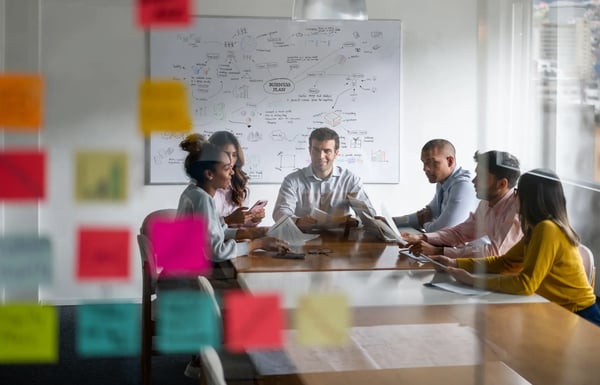 A imagem apresenta um grupo de Pessoas em uma reunião de negócios planejando sua estratégia de marketing. O tema do artigo é o que é OKR.