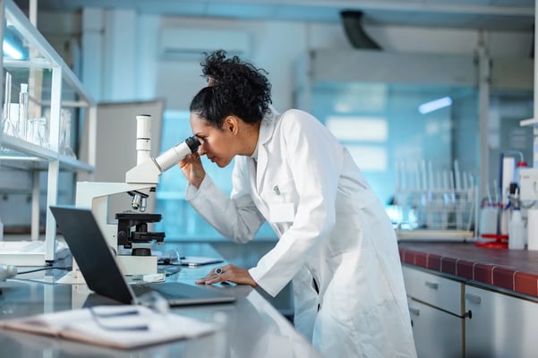 a imagem mostra uma mulher de jaleco, analisando algo em um microscópio. Ao fundo, observa-se um laboratório de análises. 