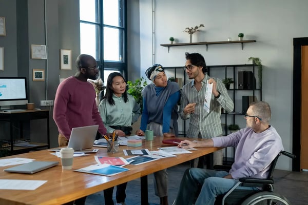 A imagem apresenta um grupo multi-étnico de pessoas trabalhando ao redor de uma mesa de reuniões, sendo que uma delas é um homem jovemde meia idade branco e cadeirante. Eles vestem roupas casuais. O tema do artigo é startups e diversidade.
