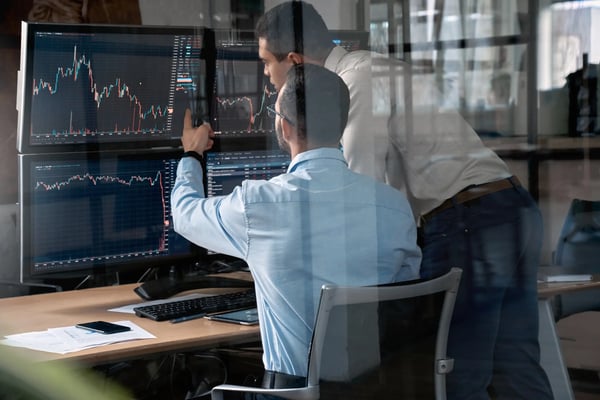 Dois homens vestindo roupa social estão em um escritório em frente a telas com gráficos de investimento
