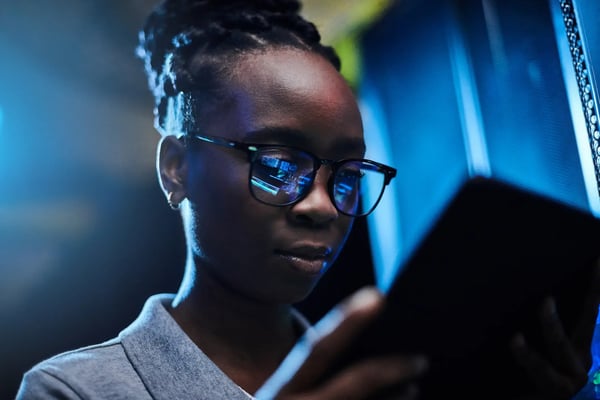 Na imagem, uma jovem mulher afro-americana em plano americano e usando óculos de grau está segurando um tablet, no qual vê algo na tela. O aparelho está de "costas" para quem vê a imagem. O fundo da imagem é escuro. O tema do artigo é tipos de inteligência artificial.