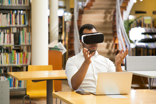 Homem usando óculos de realidade virtual gesticula sentado em frente ao computador
