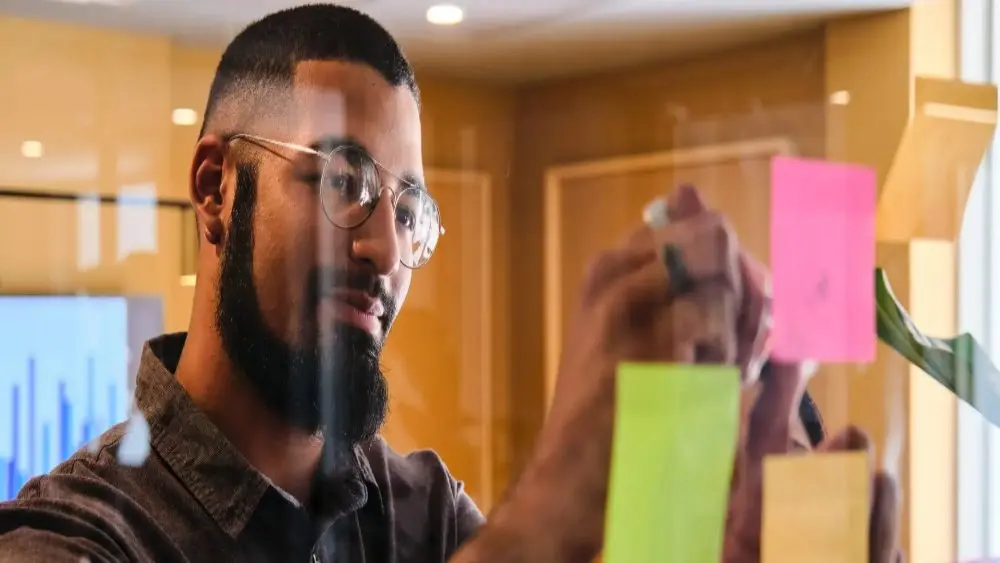 Na imagem, um homem jovem e negro, de barba e camisa marrom, usa óculos e está em frente a uma parede de vidro, colando post-its. Ele sorri. O tema do artigo é metodologias de gestão de metas.