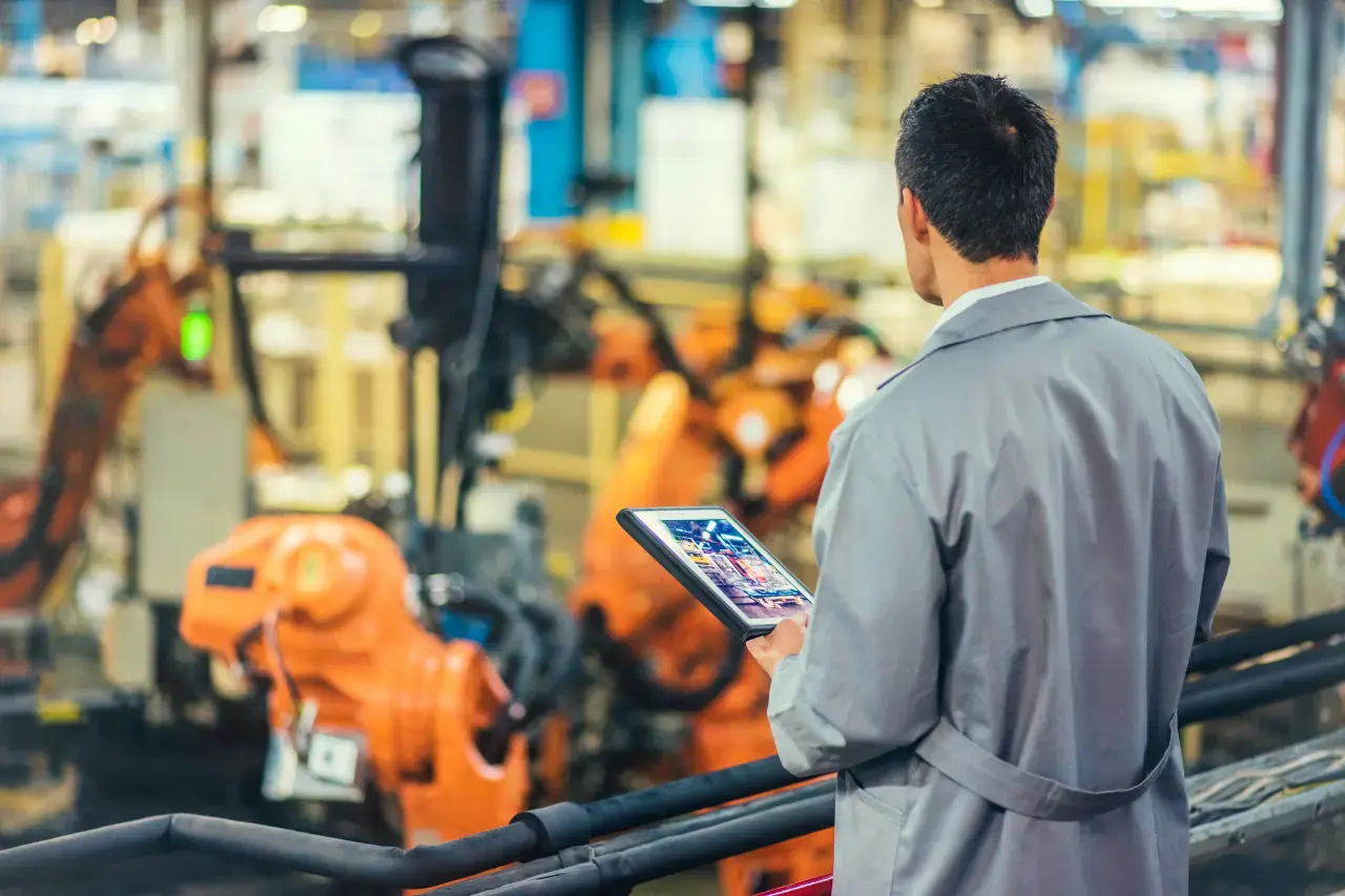 A imagem mostra um homem de costas que atua em uma fábrica operando braços mecanizados que automatizam um processo de fabricação. O tema do artigo é internet das coisas.