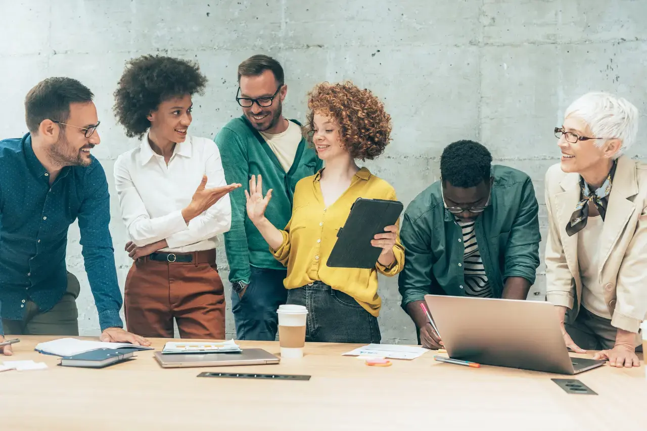 A imagem apresenta um grupo multi-étnico de pessoas em pé ao redor de uma mesa de reuniões, conversando de forma descontraída. O tema do artigo é startups e diversidade.